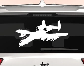 Könnte beinhalten: Ein weißes Auto mit einem weißen A-10 Warthog-Silhouettenaufkleber auf der Heckscheibe. Der Aufkleber ist mittig auf der Scheibe angebracht und von einem schwarzen Rand umgeben. Das Auto hat auch einen kleineren Aufkleber desselben Flugzeugs am unteren Teil der Heckscheibe. Der Text "DINGO" ist auf dem Stoßfänger des Autos sichtbar.