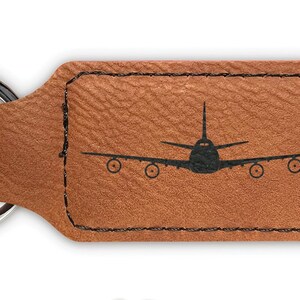 May include: Brown leather keychain with a silver key ring and a black silhouette of an airplane. The keychain is rectangular with stitched edges.