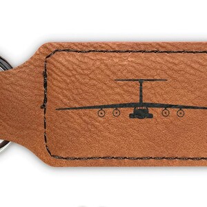 May include: A brown leather keychain with a silver key ring. The keychain features a silhouette of a C-17 Globemaster III aircraft. The keychain is available in a variety of colors.