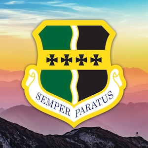 May include: A shield-shaped emblem with a yellow border, featuring green and black sections, four black crosses, and the words "Semper Paratus" in blue on a white banner. The background shows a mountain range.