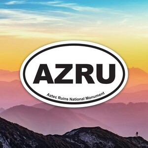 Puede incluir: Una pegatina ovalada con las letras "AZRU" en negrita negra, con "Aztec Ruins National Monument" debajo. La pegatina está colocada sobre un fondo de una cordillera y un cielo colorido.