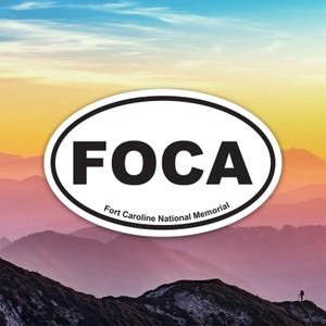 May include: An oval sticker with a black border and the word "FOCA" in bold black letters. Below the word is the text "Fort Caroline National Memorial" in smaller black letters. The background is a scenic view of mountains under a colorful sky.