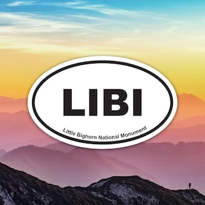 以下が含まれることがあります： 太字の黒い文字で「LIBI」という言葉が書かれた楕円形のステッカー。その下には「Little Bighorn National Monument」という文字があります。背景には、カラフルな空の下の山脈が描かれています。