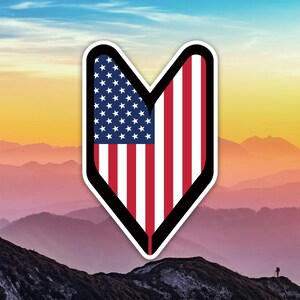 May include: A sticker featuring the American flag in a downward-pointing arrow shape. The flag design includes white stars on a blue background and red and white stripes. The sticker has a black border and is set against a mountain landscape.