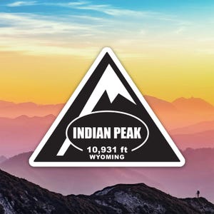 May include: A white-bordered sticker with a black triangle design, featuring a mountain graphic and the text "INDIAN PEAK" in a white oval. It also states "10,931 ft WYOMING". The background shows a mountain range under a colorful sky.