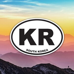 Peut inclure: Un autocollant ovale à bordure blanche présente les lettres "KR" en gras noir. En dessous, le texte "SOUTH KOREA" est imprimé en plus petit, en noir. L'arrière-plan montre une chaîne de montagnes sous un ciel coloré.