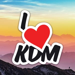 May include: A sticker with the phrase "I ❤️ KDM" in black and red. The heart is red, and the letters are black with a white outline. The background is a scenic view of mountains under a colorful sky.