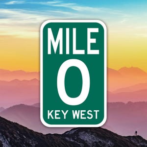 Peut inclure: Un panneau vert et blanc indiquant "MILE 0 KEY WEST" sur fond de chaîne de montagnes et de ciel coloré. Le panneau est rectangulaire avec des coins arrondis.