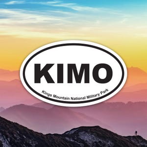 May include: An oval sticker with the word "KIMO" in bold black letters, and "Kings Mountain National Military Park" in smaller text. The sticker has a black border and is set against a mountain landscape with a colorful sky.