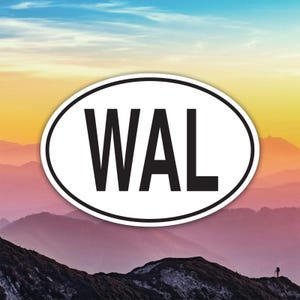 May include: Oval sticker with the letters "WAL" in bold black font, set against a backdrop of a mountain range under a colourful sky. The sticker has a white border.