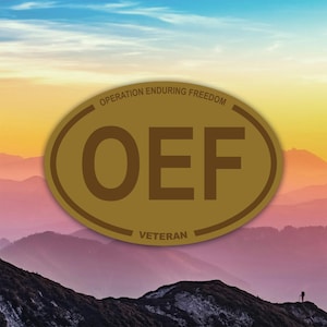 May include: A gold-colored oval emblem with the letters "OEF" in bold, dark brown font. The text "OPERATION ENDURING FREEDOM" arches above, and "VETERAN" is below. The background shows a mountain range under a colorful sky.