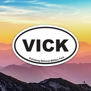 Könnte beinhalten: Ovaler Aufkleber mit dem Wort "VICK" in fetten schwarzen Buchstaben, schwarz umrandet, auf weißem Hintergrund. Der Text "Vicksburg National Military Park" steht darunter. Der Hintergrund zeigt eine Bergkette unter einem farbenfrohen Himmel.