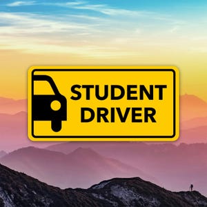 Könnte beinhalten: Ein gelbes, rechteckiges Schild mit abgerundeten Ecken, das eine schwarze Autosilhouette und den Text "STUDENT DRIVER" in fetten schwarzen Buchstaben zeigt. Der Hintergrund zeigt eine Bergkette unter einem farbenfrohen Himmel.