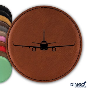 May include: A brown leather coaster with a black silhouette of an airplane in the center. The coaster has a stitched edge.