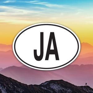 May include: White oval sticker with the letters "JA" in black, set against a scenic background of mountains and a colourful sky. The sticker has a black border.