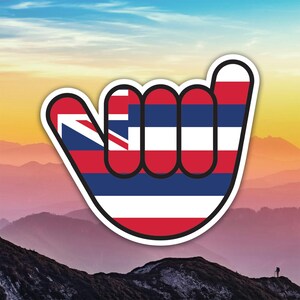 May include: A sticker featuring the shaka hand gesture, with the Hawaiian flag design. The design includes red, white, and blue stripes, and a small Union Jack in the upper left corner. The sticker has a white border and is set against a mountain landscape.
