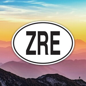 Può includere: Un adesivo ovale con le lettere "ZRE" in grassetto nero su sfondo bianco. L'adesivo è posto su uno sfondo di montagne e un cielo colorato.