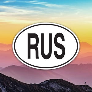 May include: An oval sticker featuring the letters "RUS" in bold black, on a white background. The sticker is set against a backdrop of mountains and a colourful sky, suggesting travel or a destination.