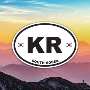 Peut inclure: Autocollant ovale blanc avec le texte noir "KR" et "SOUTH KOREA". L'autocollant a une bordure noire et de petits drapeaux sud-coréens de chaque côté de "KR". Le fond est une chaîne de montagnes avec un ciel coloré.