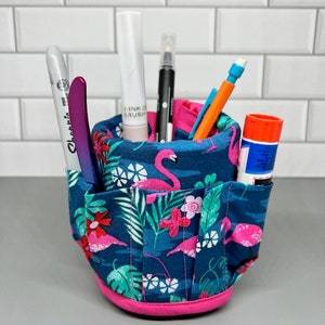 May include: A blue and pink fabric pencil holder with flamingo and tropical leaf designs. The holder has multiple compartments for organizing pens, pencils, and other supplies.