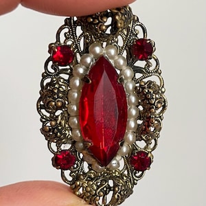 Può includere: Un pendente ornato in stile vintage con una grande gemma rossa sfaccettata al centro. La gemma è circondata da piccole perle bianche e gemme rosse più piccole. Il pendente presenta un design a filigrana con finitura in metallo dorato.