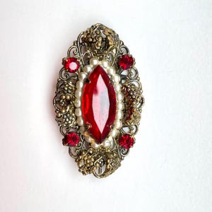 Può includere: Una spilla ovale con una cornice in metallo filigranato color oro antico. Al centro si trova una grande pietra preziosa rossa taglio marquise circondata da un anello di piccole perle bianche artificiali. Quattro pietre rosse rotonde accentuano i bordi.