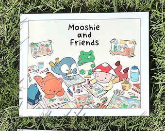 May include: Two children's books on green grass. The top book is titled "Mooshie and Friends" and features cartoon characters drawing. The bottom book has two crayon illustrations and the text "Thank You!" and "Mooshie and Friends".