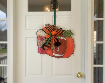 May include: A decorative fall-themed door hanger featuring a football and a coffee cup. The football is orange and brown, and the coffee cup is red. The hanger is adorned with a bow in orange, black, and green. The text on the coffee cup reads "It's the Season".