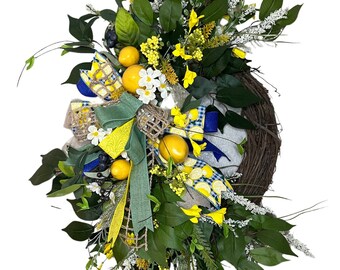 May include: A decorative wreath featuring a grapevine base, adorned with faux lemons, white daisies, and various greenery. The wreath is embellished with yellow and blue patterned ribbons and a green ribbon.