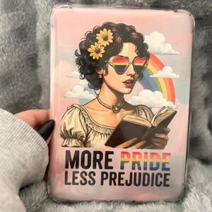 Op de afbeelding: Roze telefoonhoesje met een afbeelding van een vrouw die een boek leest, met een hartvormige zonnebril in regenboogkleuren. De achtergrond toont een regenboog en wolken, met de tekst "MORE PRIDE LESS PREJUDICE" in regenboogkleuren.