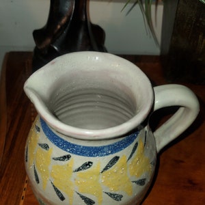 May include: A white ceramic pitcher with a yellow, blue, and black floral design. The pitcher has a handle and a wide mouth.
