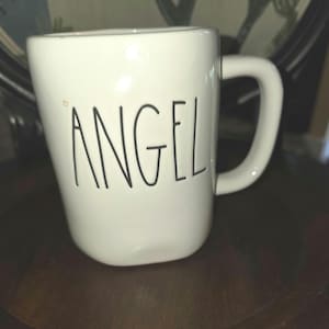 Pode incluir: Uma caneca de cerâmica branca com uma alça arredondada e uma forma ligeiramente irregular e orgânica. A palavra ANGEL está escrita em preto, com uma fonte fina de estilo manuscrito na parte frontal. A superfície é lisa e brilhante.