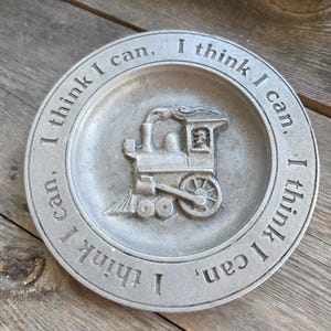 Puede incluir: Un plato redondo de color plateado con una imagen en relieve de una locomotora de vapor. La frase "I think I can" está grabada alrededor del borde. El plato está sobre una superficie de madera.
