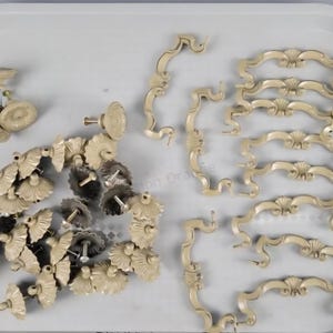 May include: A collection of cream-colored metal furniture hardware including several ornate drawer pulls with curved, decorative arms and numerous matching circular knobs featuring a central floral-inspired embossed pattern and metal mounting screws.
