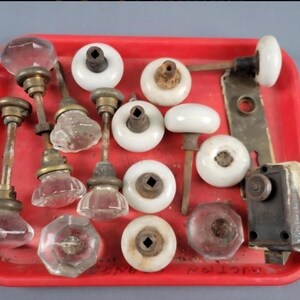 Könnte beinhalten: Eine Sammlung antiker Türknöpfe und Beschläge, darunter Knöpfe aus klarem Glas, weißer Keramik und braun, präsentiert auf einem roten Tablett. Die Beschläge weisen verschiedene Formen und Größen auf, mit Messing- und Metallstielen und Montageplatten.