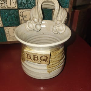 以下が含まれることがあります： 取っ手付きのセラミック製BBQ用具ホルダー。オフホワイトの容器には、中央にテクスチャ加工されたタン色のバンドがあり、長方形のラベルに「B.B.Q」の文字が書かれています。ハンドルはアーチ型で、装飾的な要素があります。