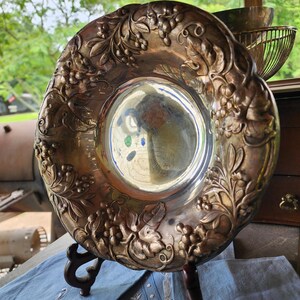 Puede incluir: Un plato decorativo plateado con un diseño en relieve de uvas y hojas alrededor del borde. El plato se exhibe sobre un soporte de madera oscura, colocado sobre una tela azul con bordados blancos. El centro del plato refleja el entorno.