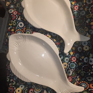 May include: Two white ceramic fish-shaped serving dishes. The dishes are on a colorful floral fabric background.