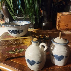 May include: A set of three blue and white ceramic kitchenware items. A small bowl with a blue heart design, a small pitcher with a blue heart design, and a small jar with a blue heart design and a cork stopper.