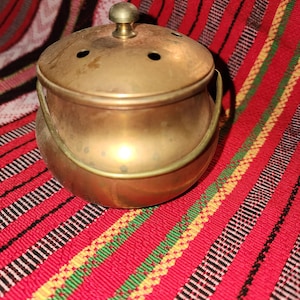 May include: A small, round, metallic brass pot with a matching lid featuring a central knob and several small ventilation holes. The pot includes a curved, hinged handle attached to the sides, showcasing a polished, warm-toned finish.