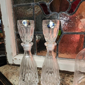 May include: Two clear glass decanters with a crystal-like design. The decanters have a small, round top and a wider base. The decanters are sitting on a brown surface.