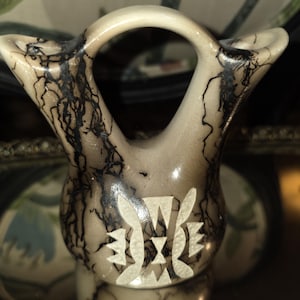 May include: A ceramic vessel with a double-spout design connected by a central arch. The surface features a smooth, light gray finish with dark, branching lightning-like patterns and a carved, white geometric emblem on the front center.