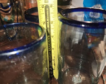 May include: Clear glass tumblers with a blue rim, approximately 5 inches tall, are shown. A yellow measuring tape is placed next to the glasses. The glasses are on a brown surface.