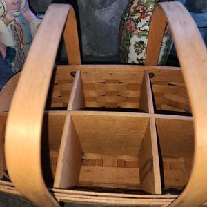May include: A light brown, woven wooden basket with a handle on each side. The interior is divided into six compartments. The basket is designed to carry bottles or other items. The handles are made of curved wood.