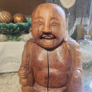 Puede incluir: Una estatua de Buda de madera con una expresión sonriente. La estatua está tallada en madera de color marrón claro y tiene un diseño detallado. El Buda está sentado y tiene una división en el medio.