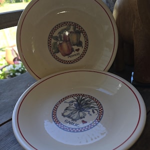 May include: Two off-white ceramic bowls with a red rim. One bowl features a pepper design with the word "PEPPERS", and the other has a garlic design with the word "GARLIC". The bowls are decorated with a blue and red dotted border.