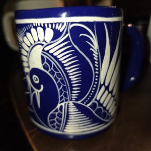 May include: A deep blue ceramic mug featuring a hand-painted white bird design with intricate line work and patterns. The mug has a cylindrical shape, a solid blue handle, and a smooth finish, showcasing a contrasting white bird motif on the side.