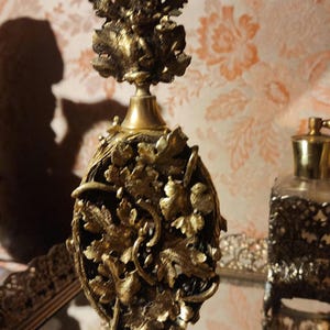 May include: Ornate antique perfume bottle with a gold finish. The bottle features intricate leaf and floral designs, with a detailed top. The background includes a floral patterned wallpaper and a mirror.