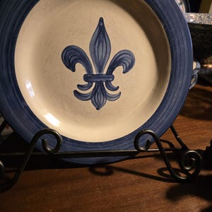 Pode incluir: Um prato de cerâmica com um centro creme e uma borda azul escuro. O prato apresenta um desenho de flor-de-lis azul escuro no centro. O prato é exibido em um suporte de prato de metal preto.