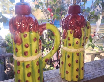 May include: Two decorative ceramic oil and vinegar dispensers. The dispensers are green with red polka dots and a red top. Each dispenser has a handle and a spout with a white stopper. The dispensers are on a wooden surface.